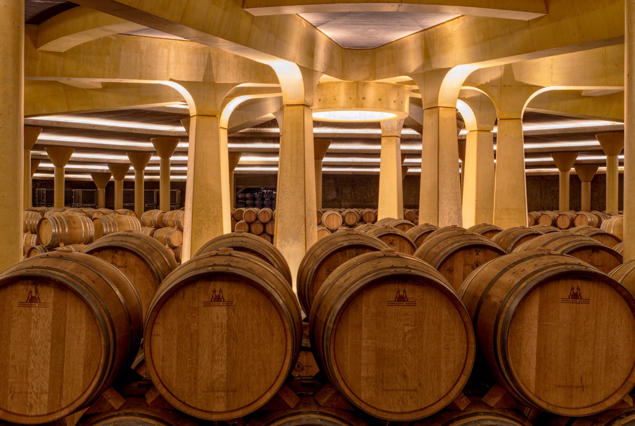 Rows of oak barrels stacked four high in a long stone cellar with candle-glow lighting