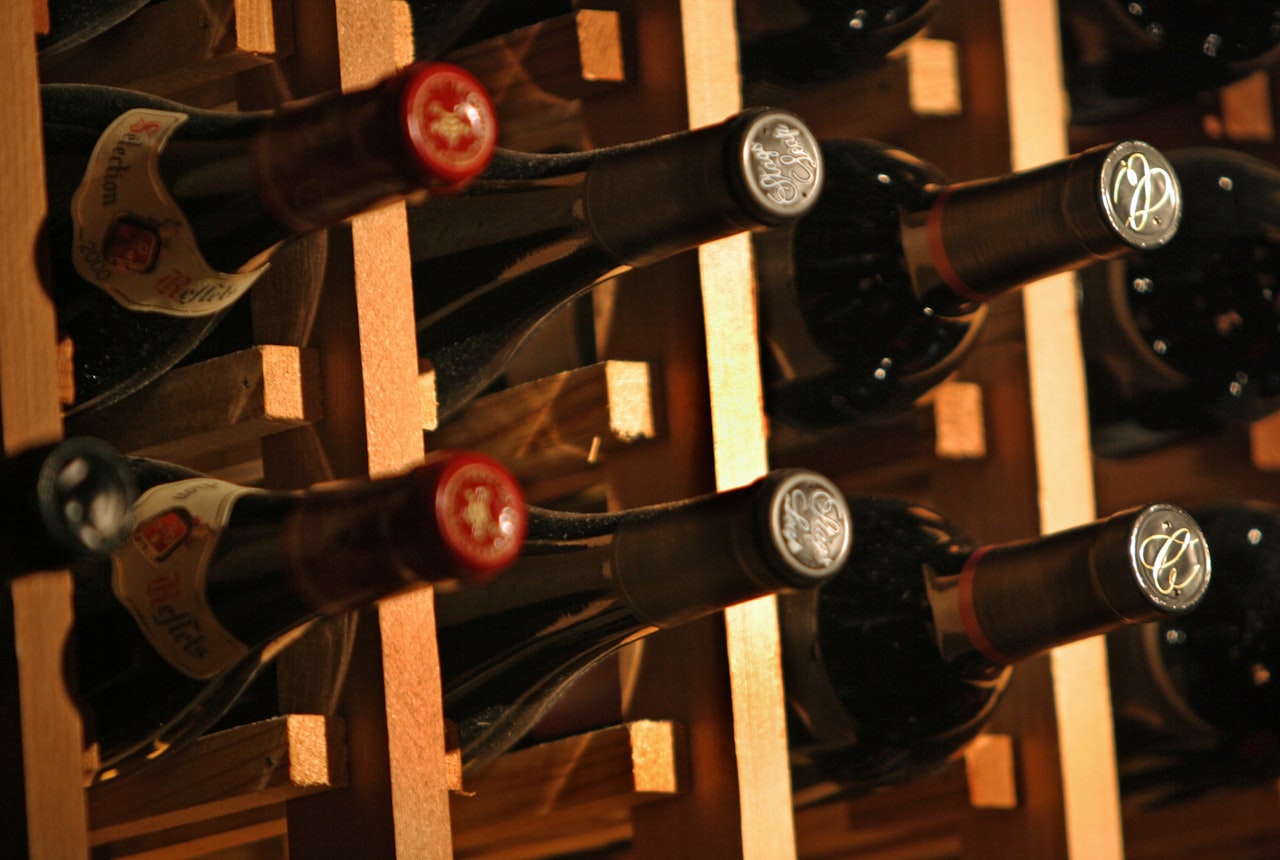 Wine bottles on a stable wooden shelf in a quiet room contrasted with the side of a refrigerator at the edge of frame